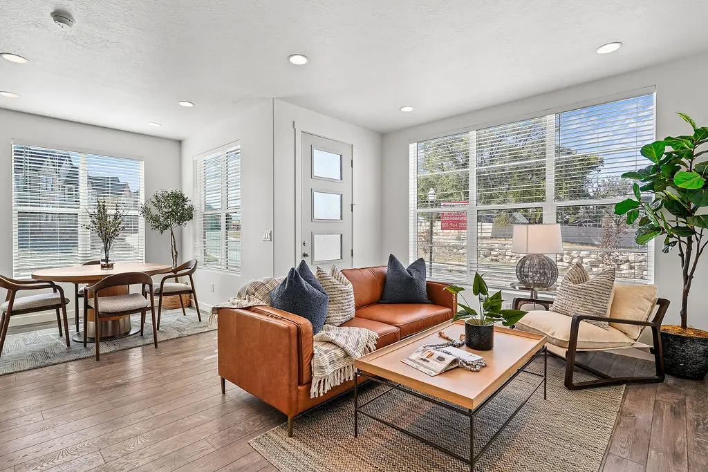 A cozy and modern living room featuring a brown leather sofa with decorative pillows, a beige armchair, a wooden coffee table with a plant, and a dining area with wooden chairs and a round table. Large windows provide natural light.