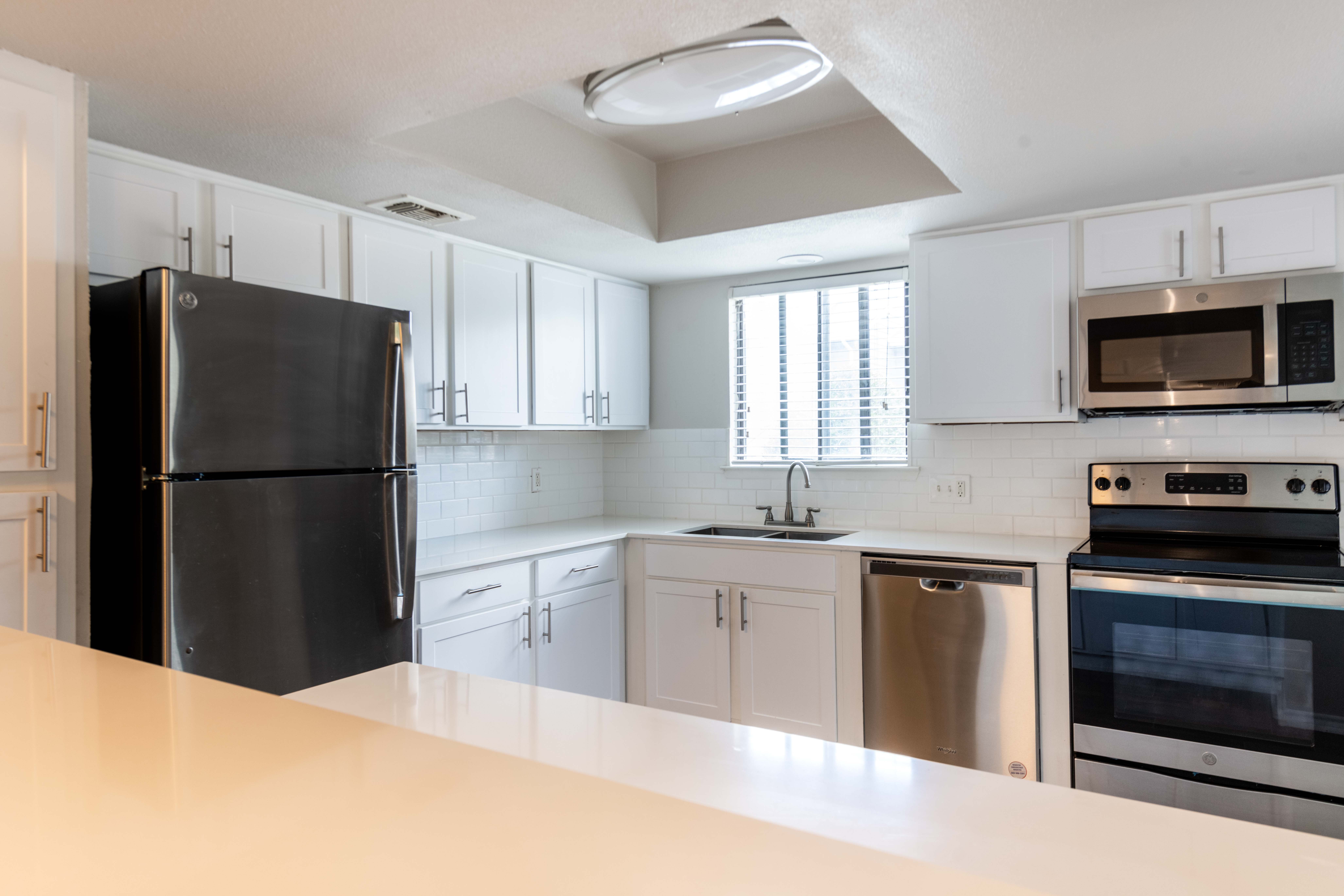 A modern kitchen featuring white cabinets, stainless steel appliances, and a bright atmosphere. The kitchen includes a refrigerator, oven, microwave, and sink, with a window allowing natural light.
