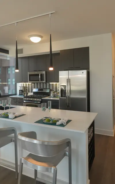 A modern kitchen featuring a large island with two barstools, stainless steel appliances, and a bright, open layout that leads into a small office area.