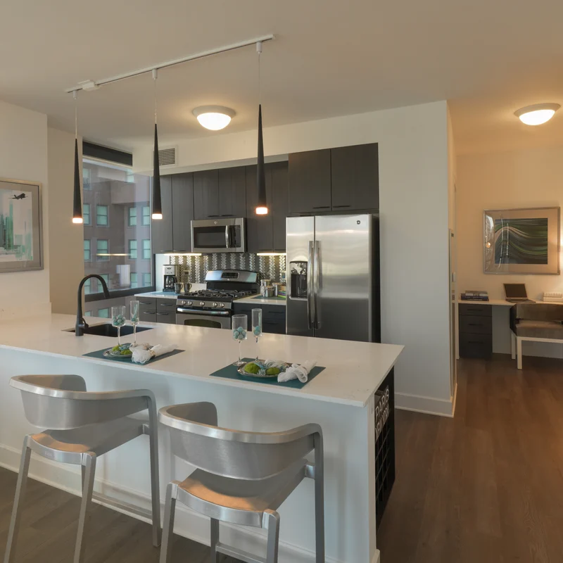 A modern kitchen featuring a large island with two barstools, stainless steel appliances, and a bright, open layout that leads into a small office area.