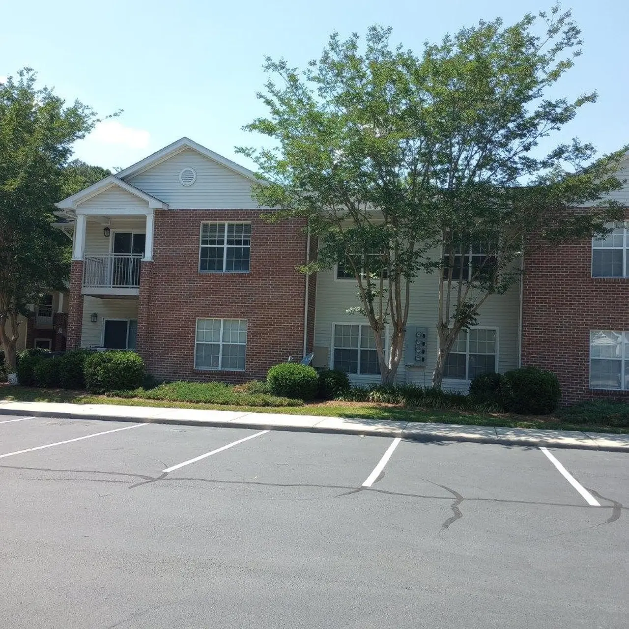 A brick apartment building with several windows and a balcony, surrounded by lush greenery and an empty parking lot.