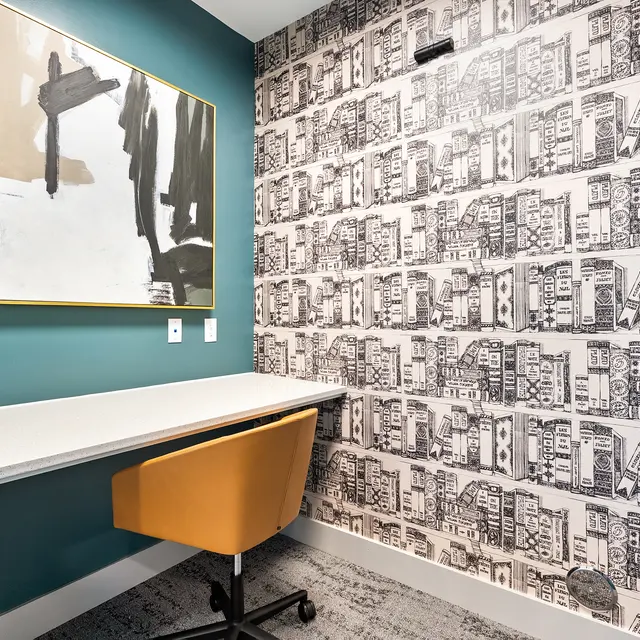 A small modern workspace featuring a white desk, a bright orange chair, and a decorative wall with black and white patterned wallpaper. An abstract painting hangs above the desk on a teal wall.