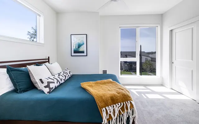 Bright and airy modern bedroom with a large window, a bed adorned with teal and patterned pillows, and a mustard throw blanket. The room features white walls and carpet, along with a stylish artwork on the wall.
