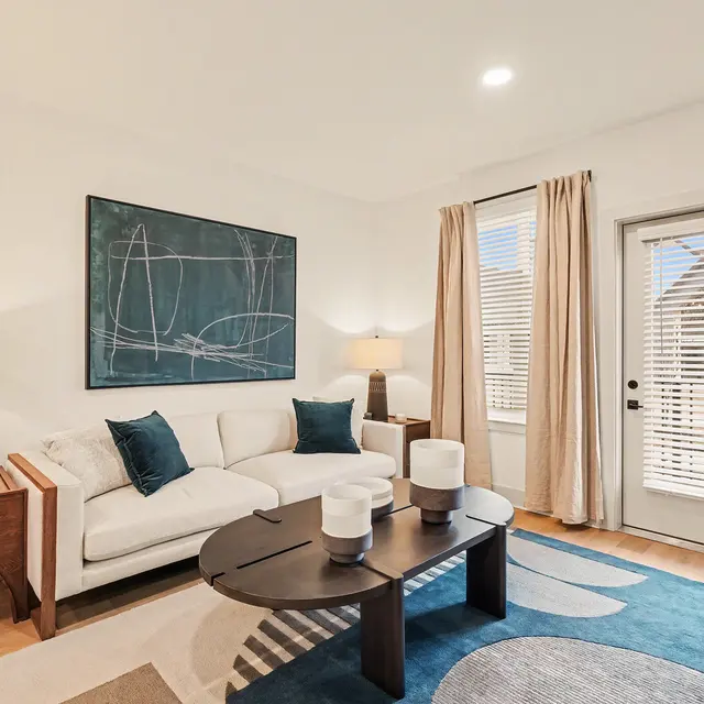 A modern living room featuring a white sofa with blue pillows, a black coffee table, and a large abstract painting on the wall. Two lamps are placed on side tables, and a door with blinds opens to an outdoor view.