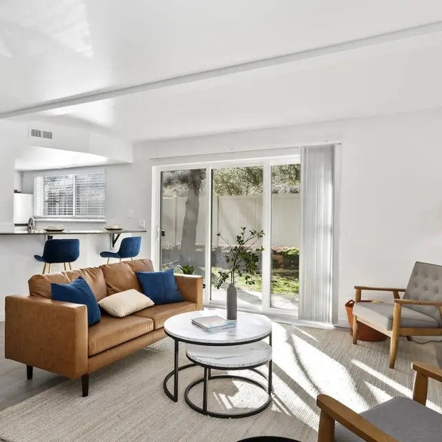 Monterra Townhomes - Modern Living Room Design A modern living room featuring a brown leather sofa with blue cushions, a round coffee table, and large sliding glass doors leading to an outdoor area. The space is brightly lit with natural light, and minimalist decor includes a floor lamp and potted plants.