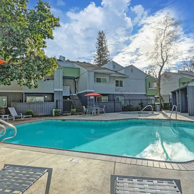 A tranquil pool area in an apartment complex featuring a clear blue swimming pool surrounded by lounge chairs and red umbrellas. The backdrop includes several buildings and trees under a partly cloudy sky.