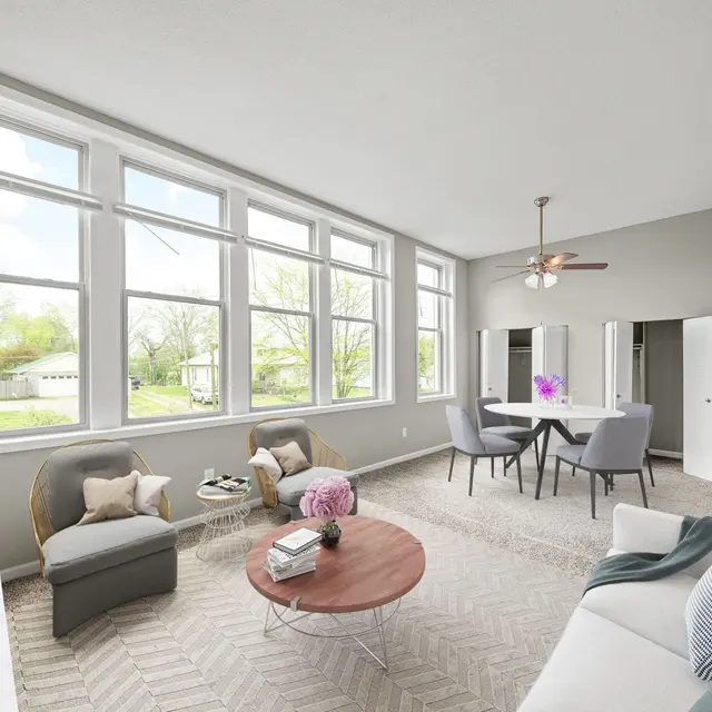 A modern living room with large windows, a round coffee table in the center, and a dining area with a table and chairs. The room is decorated in neutral tones with a mix of contemporary furniture.