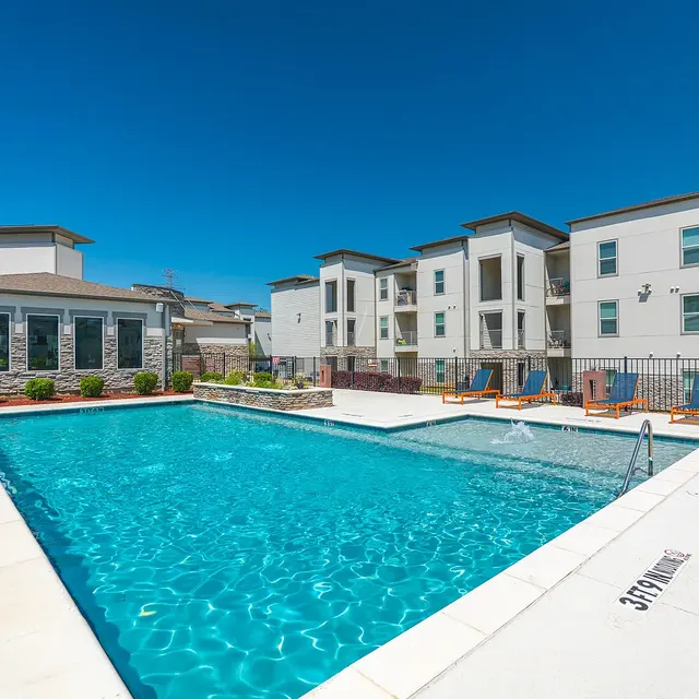 A bright outdoor swimming pool area with a clear blue sky and modern apartment buildings in the background.