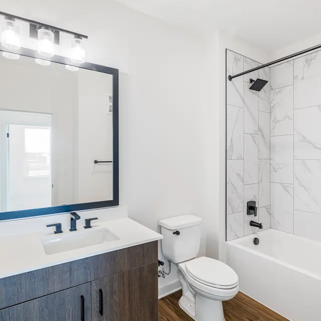 A modern bathroom featuring a large mirror above a sleek vanity with dark wood cabinetry. A white bathtub with a black faucet is visible, along with a tiled shower wall. The space has natural light coming from a door in the background.
