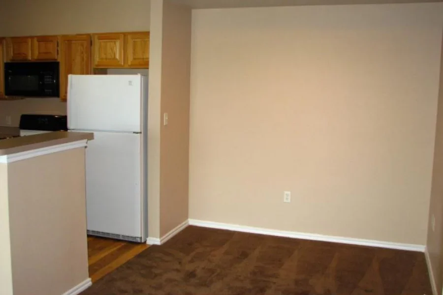 An empty living space featuring a kitchen area with wooden cabinets, a white refrigerator, and a spacious carpeted area on the right side.