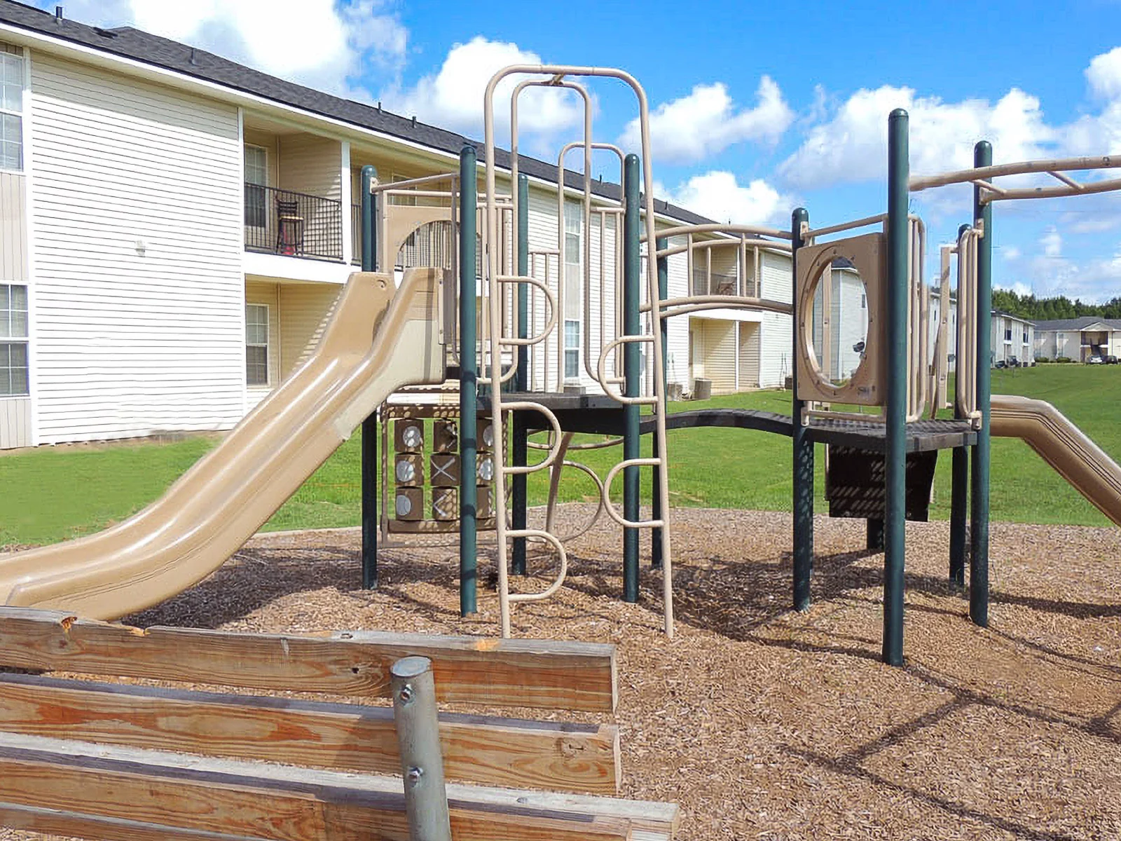 A playground featuring a slide, climbing structure, and seating area located near apartment buildings.