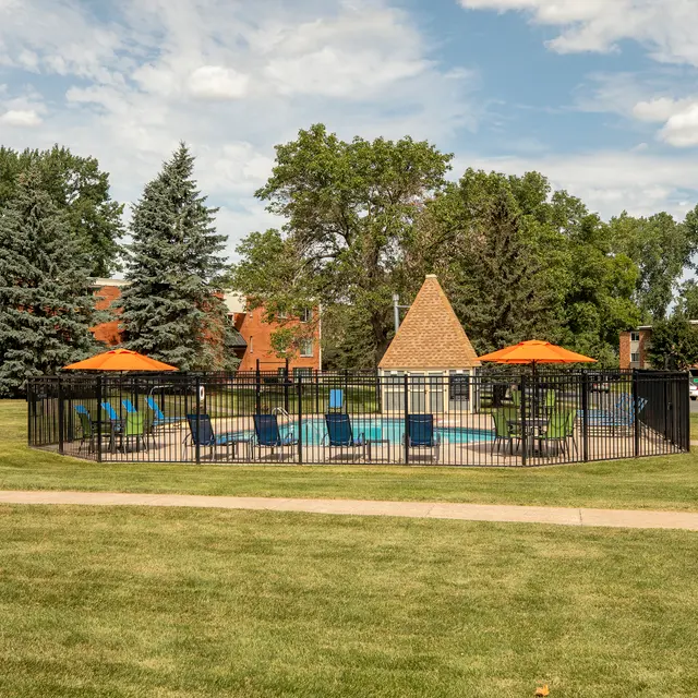 Regency Park Apartments Regency Park - A landscaped community area featuring a fenced swimming pool surrounded by lounge chairs and orange umbrellas, with residential buildings in the background and green trees.