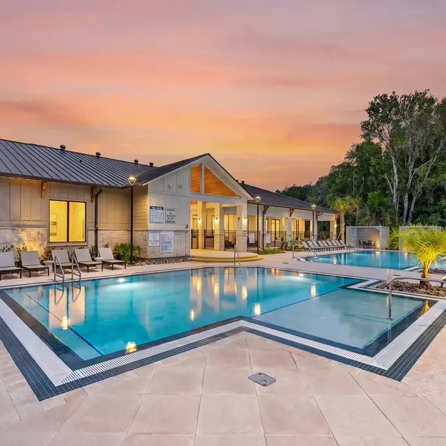 Resort-style Pool Area at Sunset A luxurious outdoor pool area featuring two swimming pools, surrounded by lounge chairs and greenery, with a modern building in the background and a colorful sunset sky.