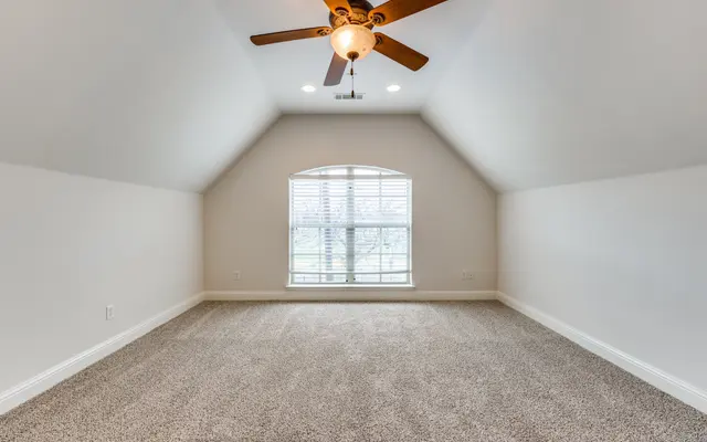 Spacious Attic Room A spacious attic room with carpet flooring, a ceiling fan, and a large window with white blinds that lets in natural light.