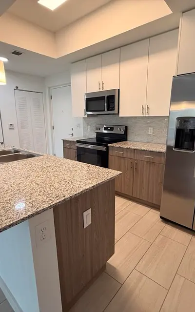 A modern kitchen with granite countertops, stainless steel appliances, and wooden cabinetry. The kitchen features an island with a sink and pendant lighting.