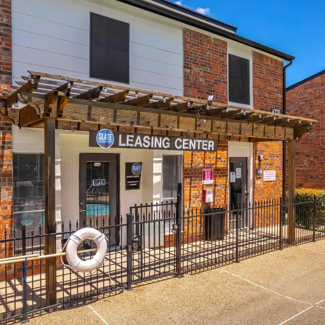 Exterior view of a leasing center with a wooden pergola and a pool float decoration, surrounded by a fence and greenery.