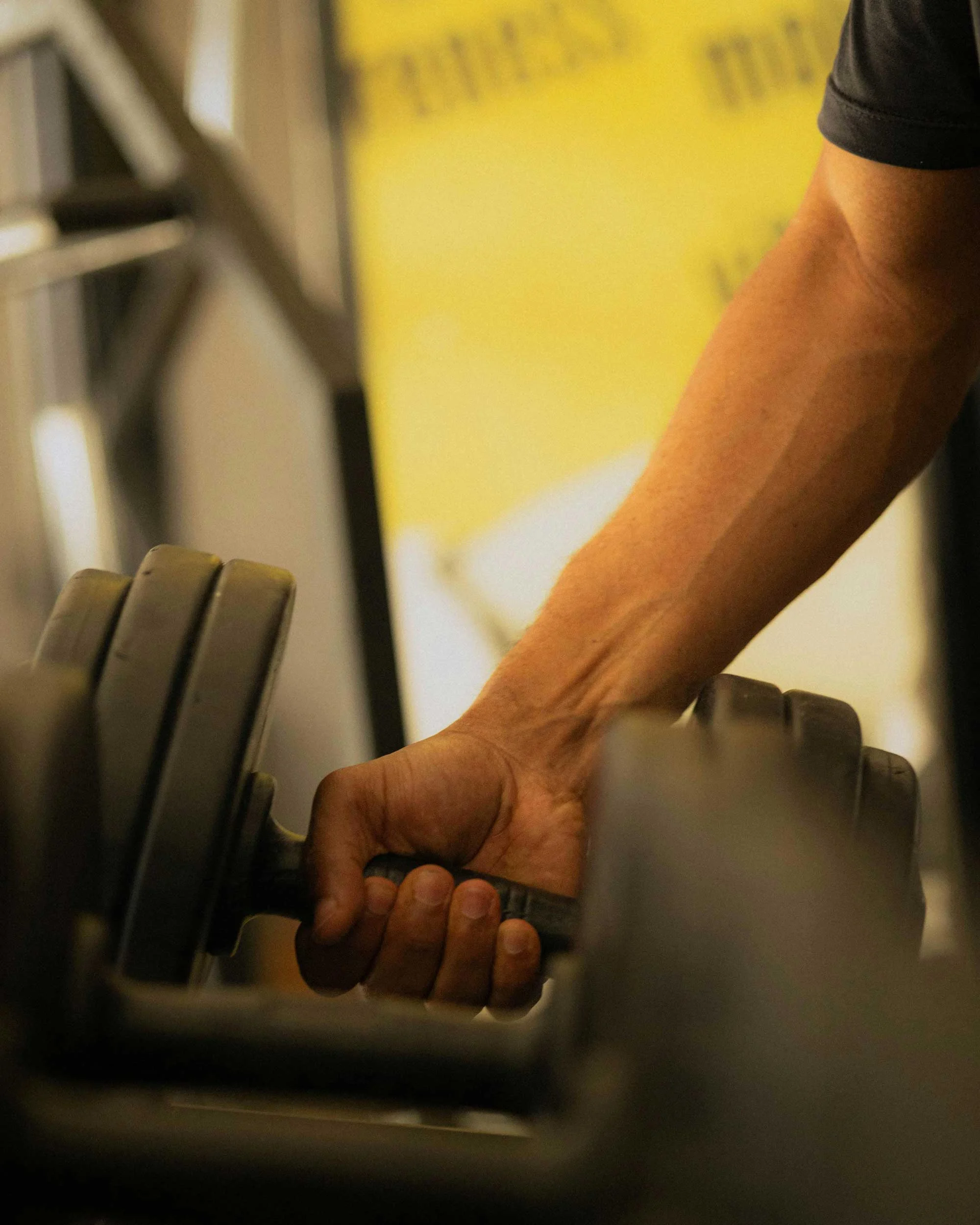 A close-up of a hand gripping a heavy dumbbell in a gym setting, with weights in soft focus in the foreground.
