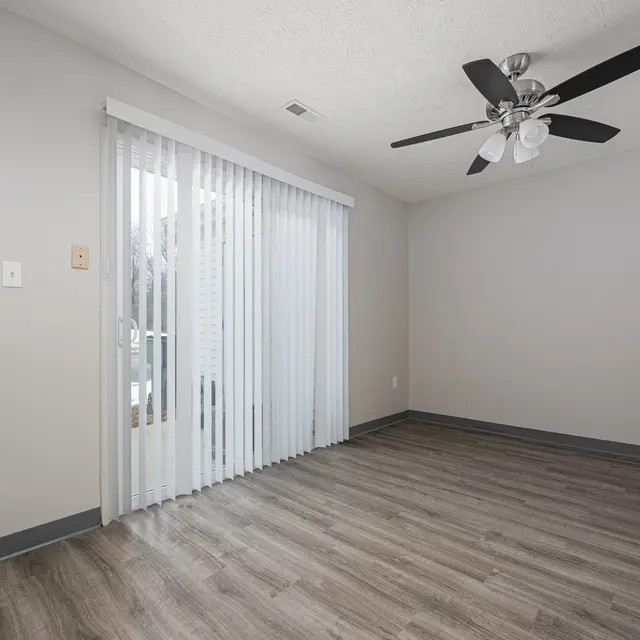 An empty room with a ceiling fan, light-colored walls, and wood-like flooring. A window with vertical blinds is visible in the background.