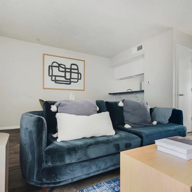 A cozy modern living room featuring a blue velvet sofa with multiple cushions. There's a small wooden coffee table with books, a lamp, and an abstract art piece on the wall. A doorway leads to another room.