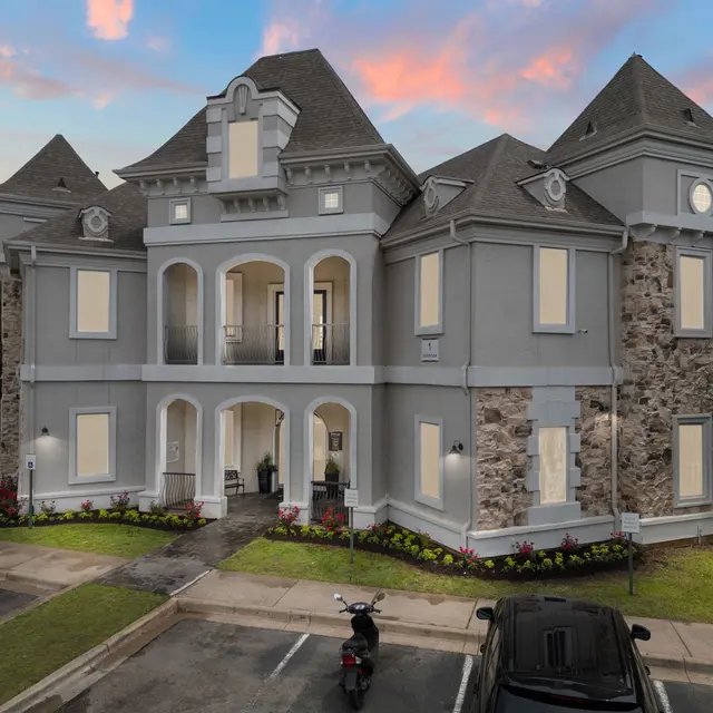 A Victorian-style mansion with gray paint and stone accents, featuring multiple gabled roofs, arched windows, and lush landscaping in the foreground