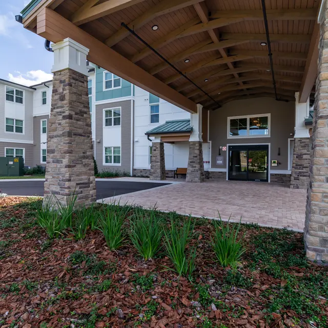 The entrance of a modern building with stone pillars and a wooden canopy, featuring a paved area and landscaped greenery in front.