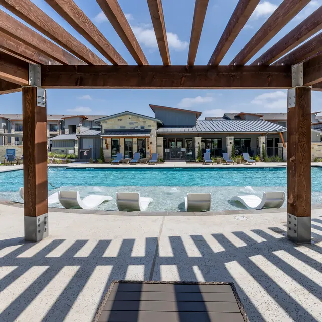 View of a swimming pool surrounded by lounge chairs and apartment buildings beneath a pergola.