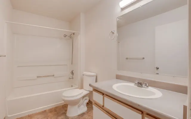 A simple bathroom featuring a bathtub, toilet, and a sink with a mirror above. The walls are painted white and the flooring has a light pattern.