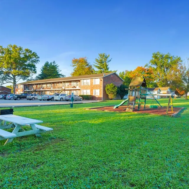 A spacious green area with a picnic table and a playground set, located near two-story apartment buildings under a clear blue sky.