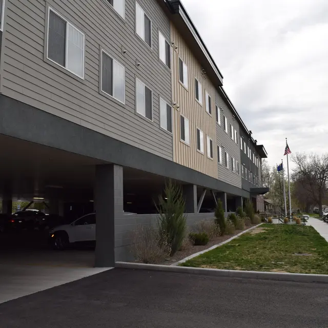 Modern Building Exterior with Covered Parking A modern building with two-tone siding and a covered parking area. The exterior features multiple windows and a sidewalk leading to the road. Trees and shrubs are visible in the grassy area alongside the parking lot.
