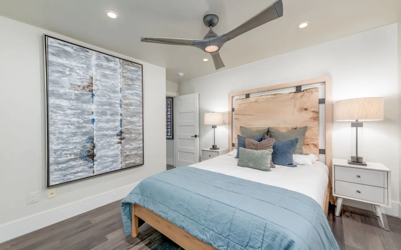 A modern bedroom featuring a stylish bed with a wooden headboard, blue bedding, and decorative pillows. Two nightstands with lamps are on either side, and an abstract artwork is hung on the wall.