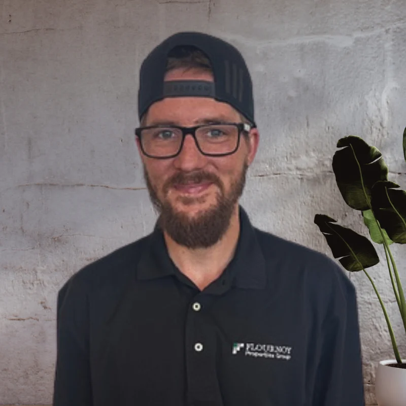 A man in a black polo shirt with glasses and a hat stands in front of a plain wall, smiling. A green plant is visible in the foreground to the right.