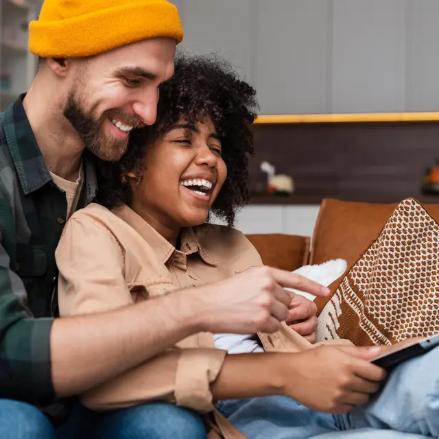 A happy couple sitting on a couch, the man in a yellow beanie and plaid shirt is playfully pointing at a tablet held by the woman with curly hair, who is laughing joyfully. They are surrounded by a cozy living room setting with a decorative pillow and some plants in the background.