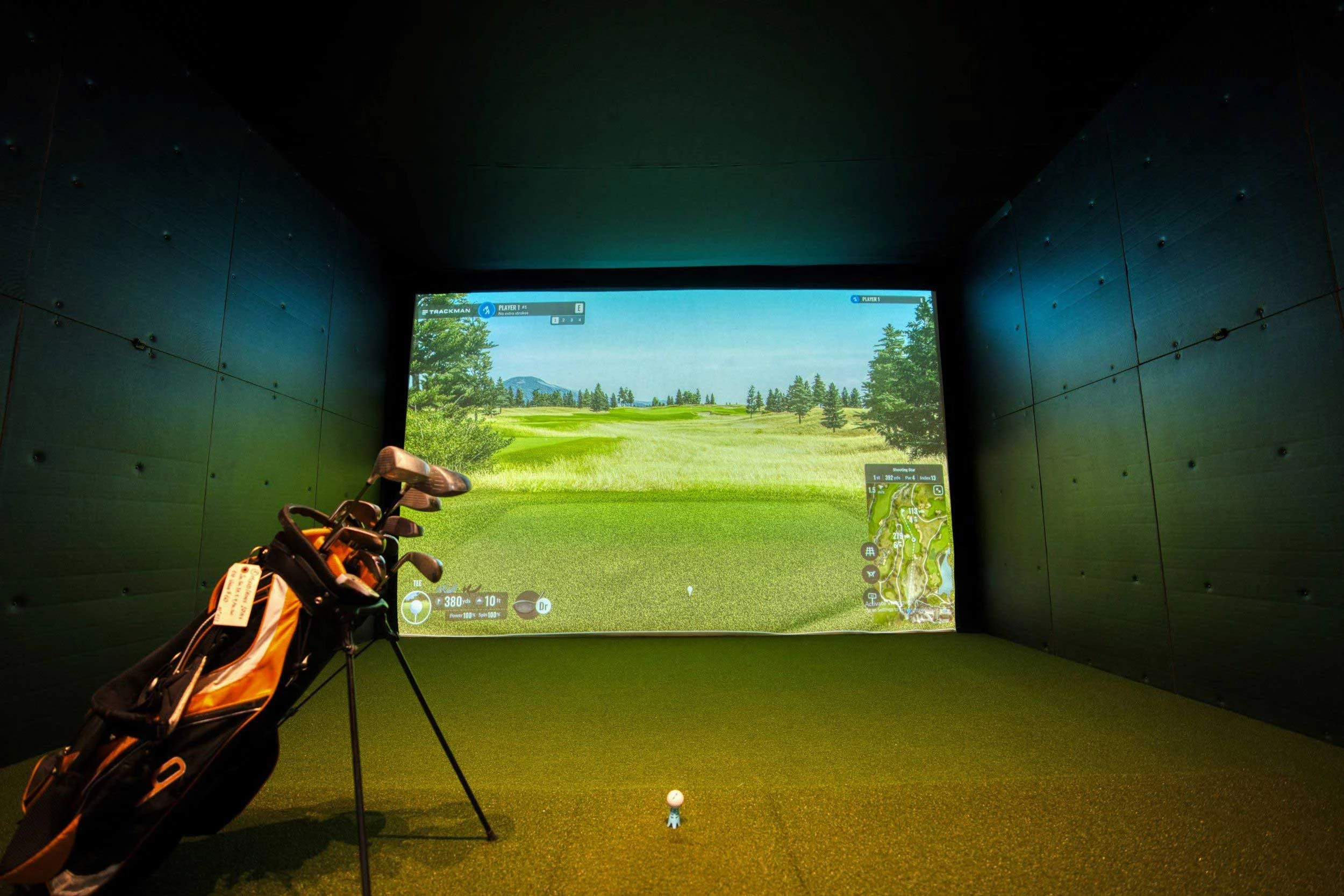 An indoor golf simulator setup featuring a golf bag in the foreground and a large screen displaying a scenic golf course.