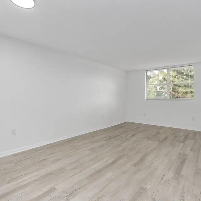 A spacious, unfurnished room with light wood laminate flooring and a large window allowing natural light to enter. The walls are painted white, contributing to a bright and open feel.