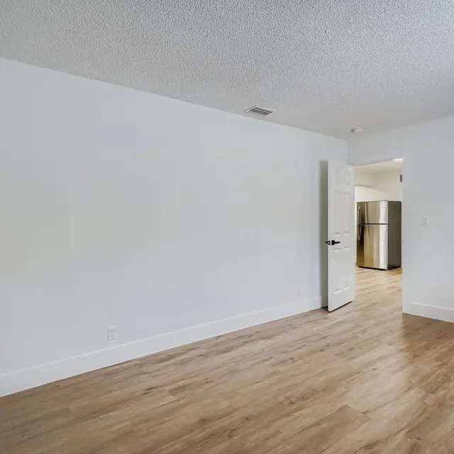 A spacious empty room with light wood flooring and a white textured ceiling. The room is partially illuminated and has a door leading to a kitchen area visible in the background. There is a doorway on the right leading to another space. The walls are painted white, creating a bright, open atmosphere.