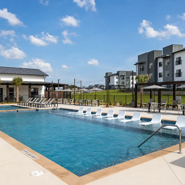 A modern swimming pool area with a clear blue sky and clouds. The pool features lounge chairs, palm trees, and a nearby building with multiple apartments.