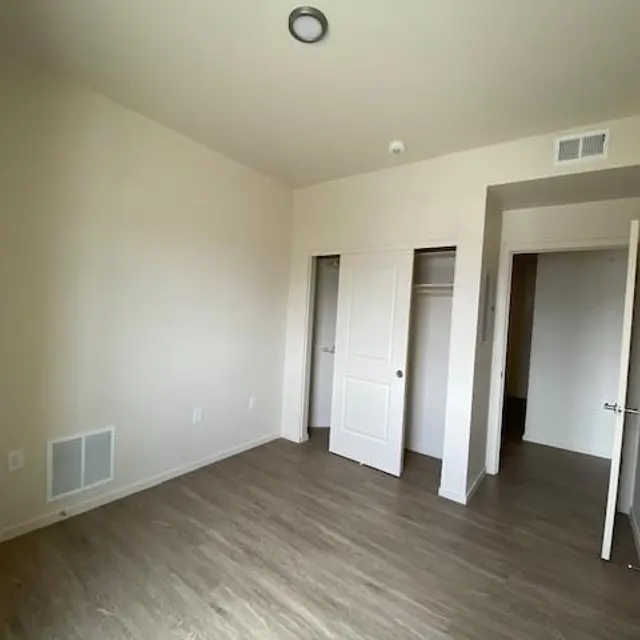 An empty room with light brown wooden flooring and light-colored walls. It has two open doors leading to closets and a ceiling light fixture.
