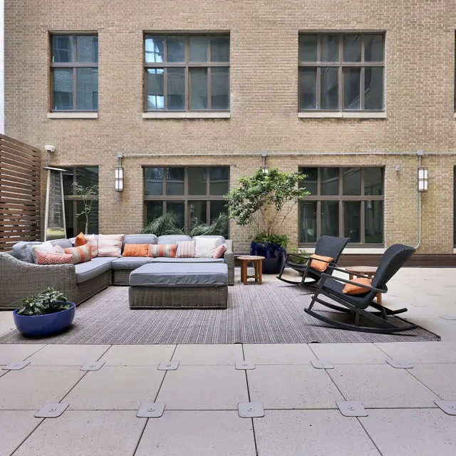 Stylish Outdoor Lounge Area A stylish outdoor lounge area featuring a gray sectional sofa with orange and white cushions, a wooden coffee table, and two rocking chairs on a tiled floor, surrounded by large windows and a small tree.