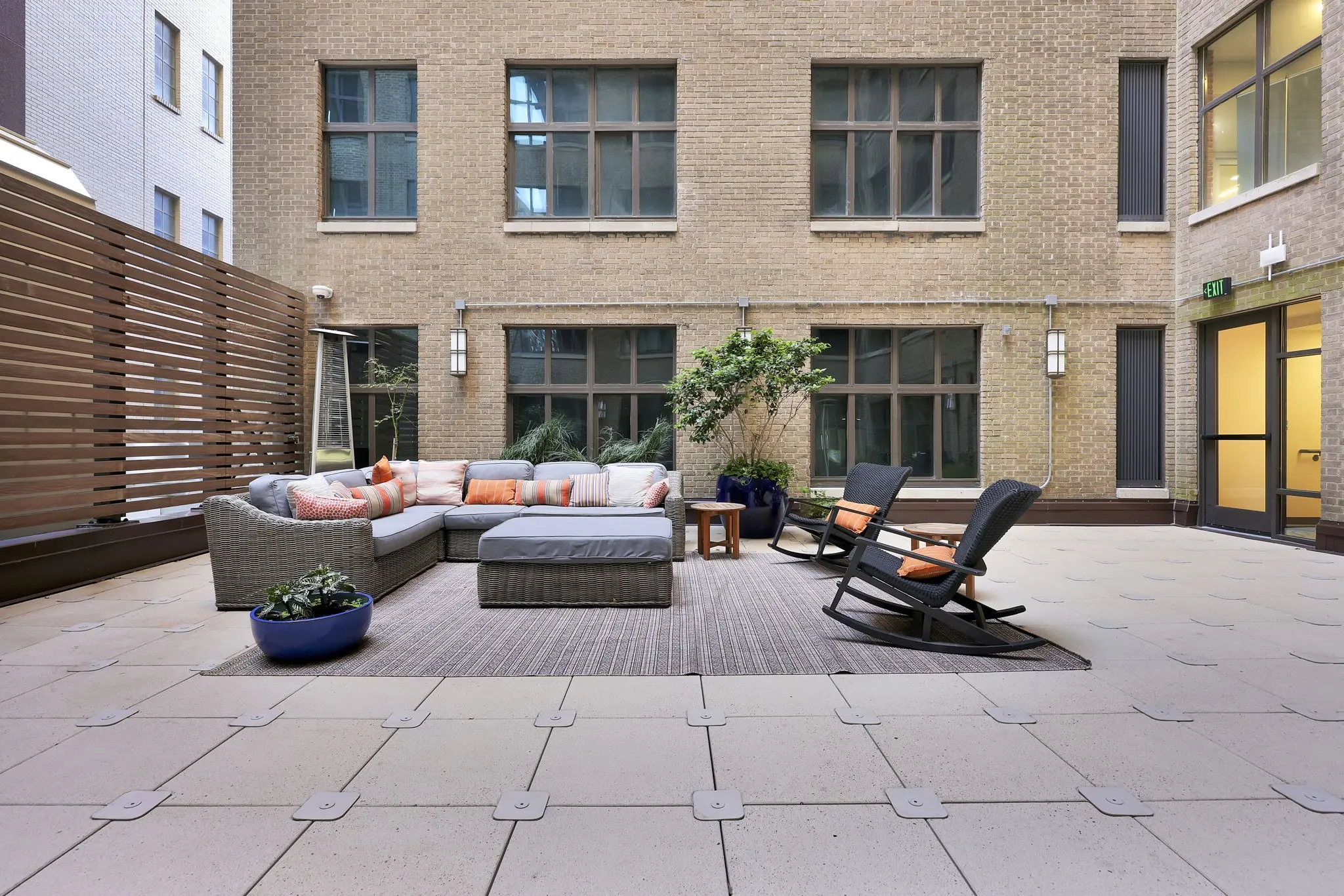 A stylish outdoor lounge area featuring a gray sectional sofa with orange and white cushions, a wooden coffee table, and two rocking chairs on a tiled floor, surrounded by large windows and a small tree.