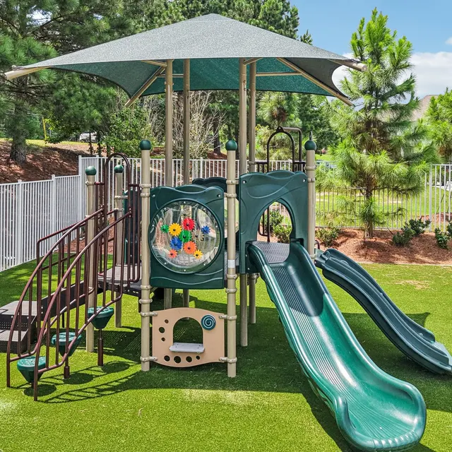 A playground featuring a slide, climbing stairs, and interactive panels, situated on green turf surrounded by trees and a fence.