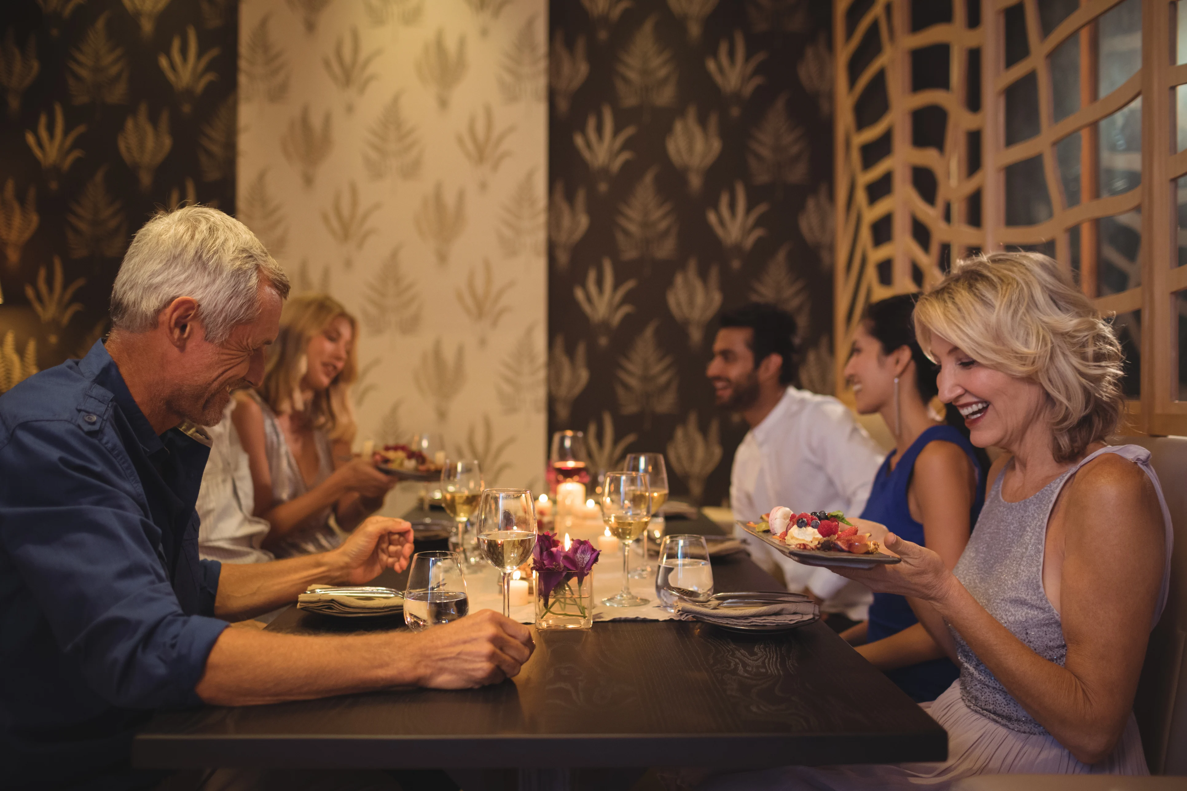 A group of five people enjoying a dinner together at a restaurant. A mix of men and women are seated at a table adorned with plates, glasses, and a decorative centerpiece. They appear to be engaged in lively conversation and laughter, with a warm ambiance created by soft lighting.