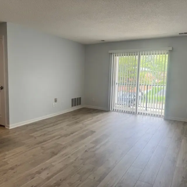 A bright living room with light gray walls and wooden floors. There are sliding glass doors leading to a balcony area, and a small kitchen area visible in the corner.