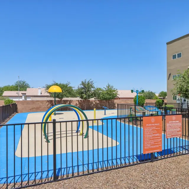 A colorful playground area enclosed by a fence, featuring a water play structure and a variety of play surfaces.
