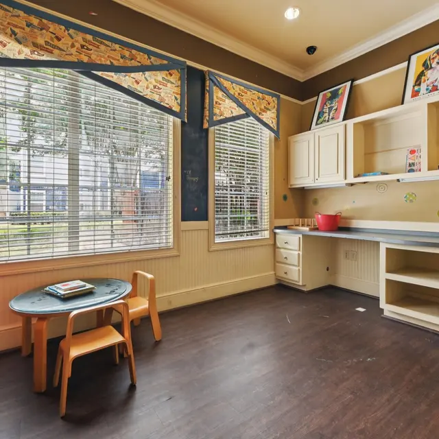 A brightly decorated playroom featuring a small table with two chairs, large windows with blinds, and built-in shelving.