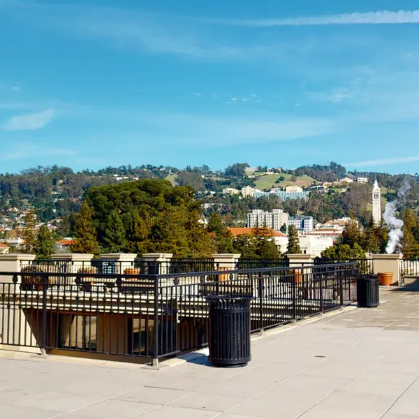 A panoramic view from a rooftop featuring a city skyline with buildings, trees, and hills in the background under a clear blue sky.