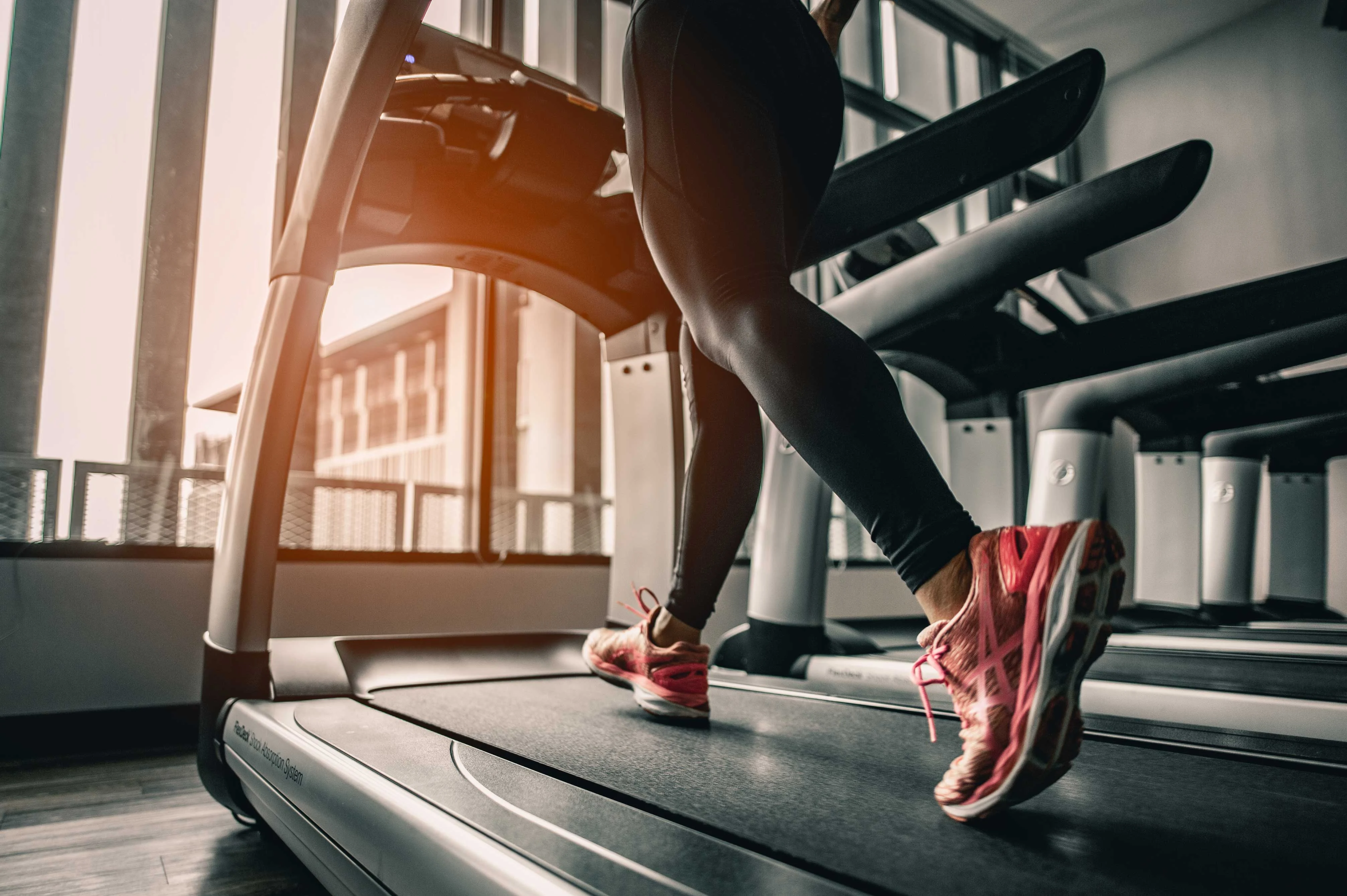 Treadmill Workout in the Gym A person running on a treadmill in a gym setting, wearing black leggings and pink running shoes, with sunlight streaming through large windows.