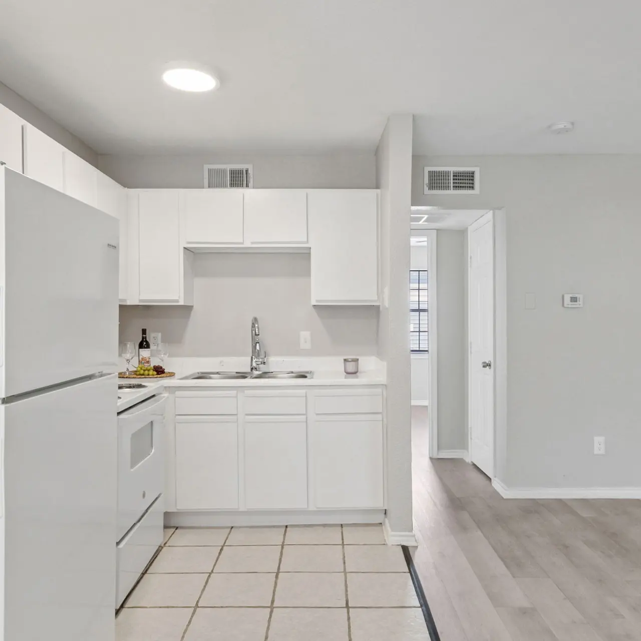 A modern kitchen featuring white cabinets, a refrigerator, and a tiled floor with an open layout leading to another room.