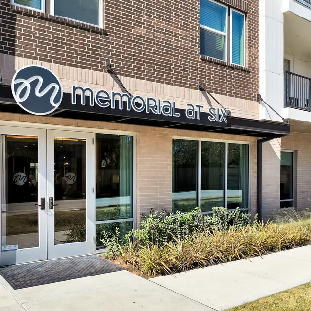 Facade of a modern building with the name 'Memorial at Six' displayed above the entrance, featuring glass doors and adjacent windows.