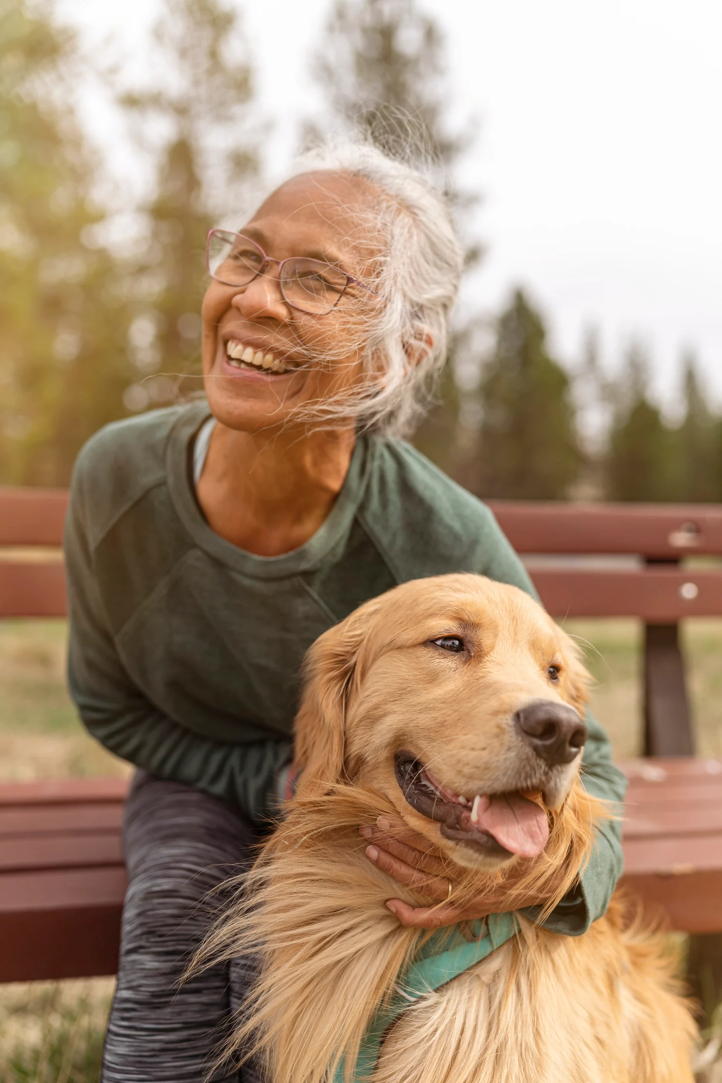 A joyful older woman with gray hair and glasses is sitting on a bench, smiling at her golden retriever dog, which has its tongue out, enjoying the moment together. They are surrounded by a natural setting with trees in the background.