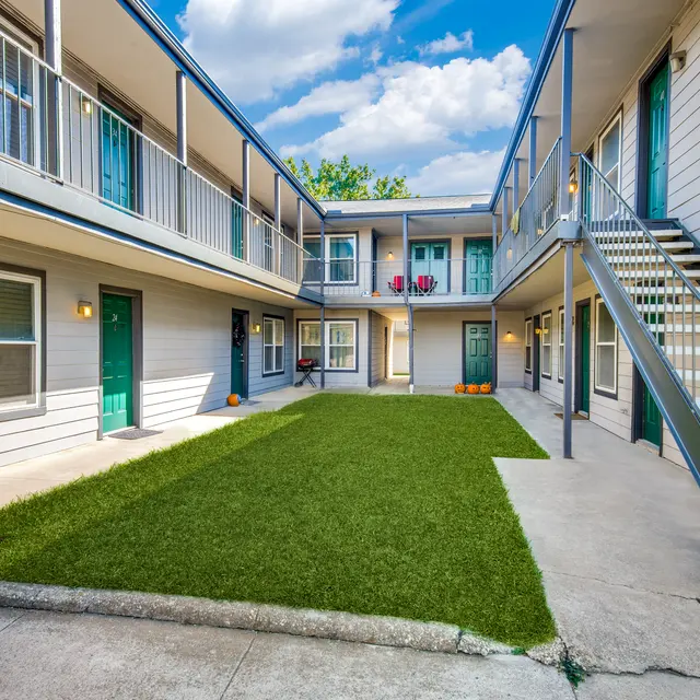 A well-lit courtyard of an apartment complex featuring green artificial grass, surrounded by two-story buildings with balconies and stairs.